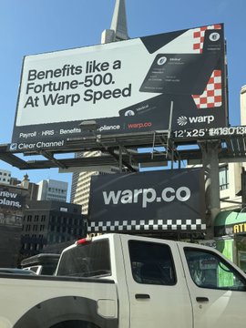 A billboard advertisement for 'warp.co' displaying 'Benefits like a Fortune-500. At Warp Speed.' above a white pickup truck on a city street.