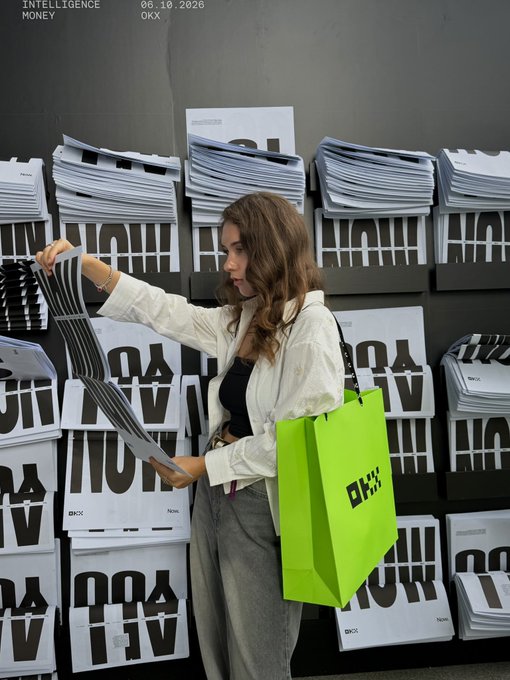 A young woman with long brown hair browses through stacks of white printed materials, carrying a bright neon green shopping bag with 'OKX' branding in what appears to be a promotional event space.