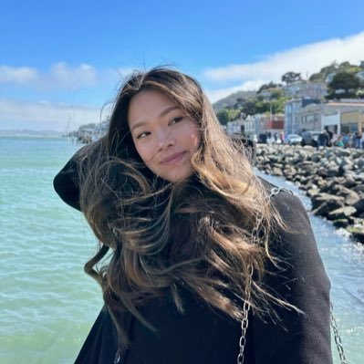 A selfie of a woman with long brown hair wearing a black top, taken outdoors with the ocean and a rocky coastline visible in the background.