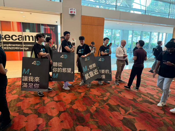 People at the TOKEN2049 conference holding black signs with the text '4M' and Chinese characters translating to 'Listening to your desires, M, let me satisfy you' and 'What you want, M, is close', promoting the 4M token.