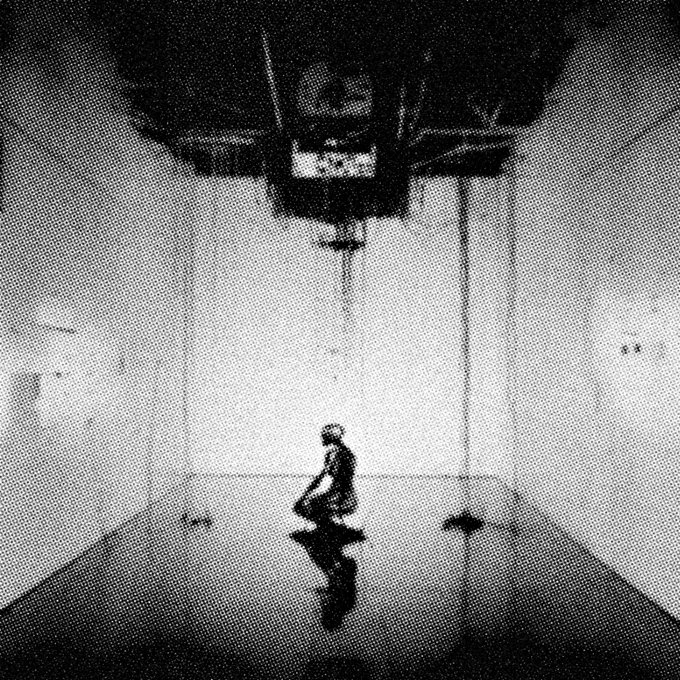 A solitary silhouetted figure stands on a reflective floor beneath a large, dark mechanical apparatus in a stark, empty room. The grainy, monochromatic composition evokes themes of isolation and experimental structure, aligning with the post's title "Uncaused Structure Event 1/1 8," which references a single-edition generative NFT on the Tezos blockchain (objkt.com).