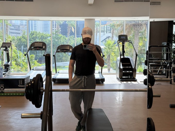 A man in a black shirt and gray sweatpants takes a mirror selfie at a gym, standing in front of a barbell with weight plates. A treadmill behind him displays "30," and his cap appears to read "Bali Bar"—visually playing on the "barbell strategy" reference to heavy weightlifting in the post's crypto investment metaphor.