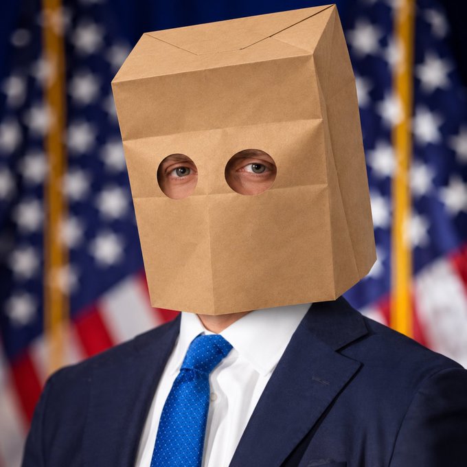 A man in a navy suit and blue tie wears a brown paper bag over his head with holes cut out for his eyes, standing in front of American flags.