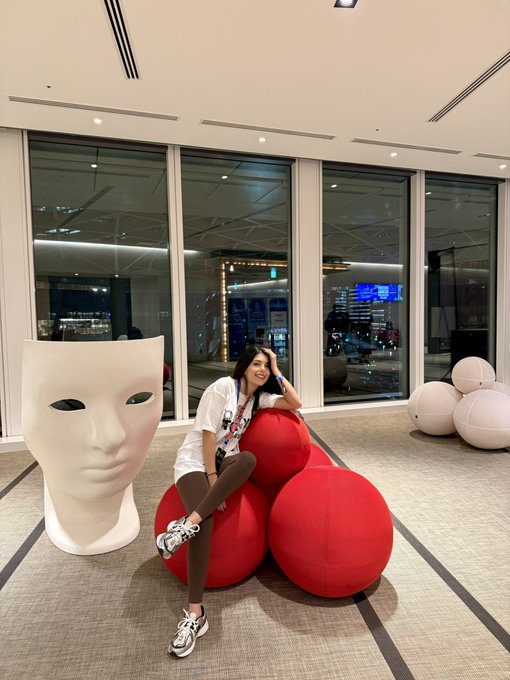 A woman poses seated on large red spherical decorations in a modern indoor space next to a large white sculptural face mask.