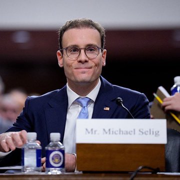 A man in a dark suit and glasses sits at a hearing table with a nameplate reading 'Mr. Michael Selig,' smiling slightly before a microphone.