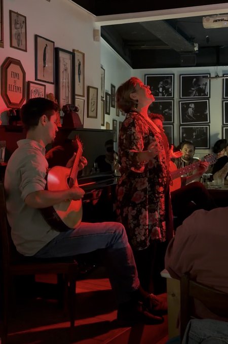 The image shows a man playing guitar while another person sings along with him. They are both dressed casually and appear to be enjoying themselves as they perform together. The room has several framed pictures hanging on the walls, adding an artistic touch to the space. In addition to the musicians, there is also a group of people sitting at tables in the background, possibly engaged in conversation or other activities.