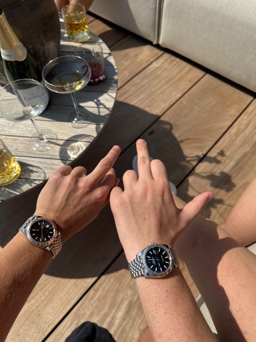 Two hands making middle finger gestures while wearing Rolex watches, sitting at an outdoor table with wine glasses and a bottle on a wooden deck.