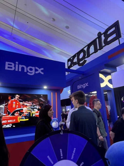 A BingX promotional booth at an event featuring Formula 1 imagery, with attendees gathered around a spinning prize wheel and branded displays.
