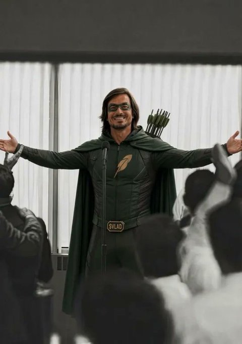 A man dressed in a green hooded costume with a quiver of arrows, resembling a TV archer character, stands with arms outstretched before a crowd with a microphone and a 'SYLAD' badge.