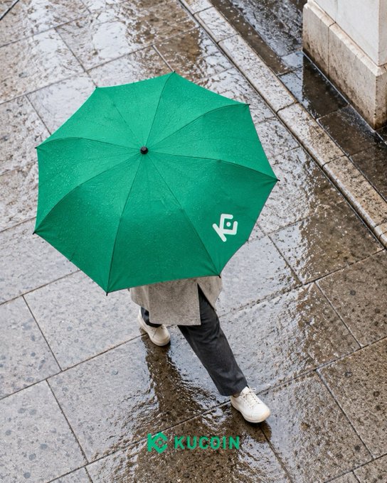 A person walks through a rain-soaked street beneath a bright green KuCoin-branded umbrella, visually echoing the post's message of weathering the storm with portfolio protection. The image cleverly ties KuCoin's safeguarding role to the literal shelter an umbrella provides on a rainy day, with the watermark "KUCOIN" displayed at the bottom.
