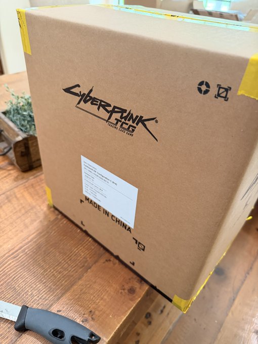 A cardboard box with the 'Cyberpunk 2077: The Board Game' logo and 'Made in China' text sits on a wooden surface next to a knife and a small plant.