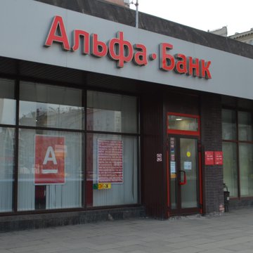 The image shows the storefront of Alfa-Bank (Альфа·Банк), with its red signage and logo visible above the entrance, along with promotional posters in the window. The image illustrates the bank referenced in the post, which is reportedly piloting cryptocurrency trading through its Alfa-Investments app for qualified investors.