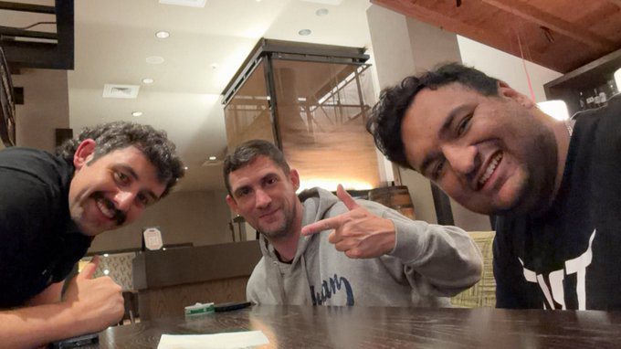 Three smiling men pose together at a table in a modern interior with wood-beamed ceilings, with the man on the right wearing a black shirt featuring a white "87" logo. The man in the center wears a gray hoodie with partially visible text, while the man on the left gives a thumbs-up gesture.