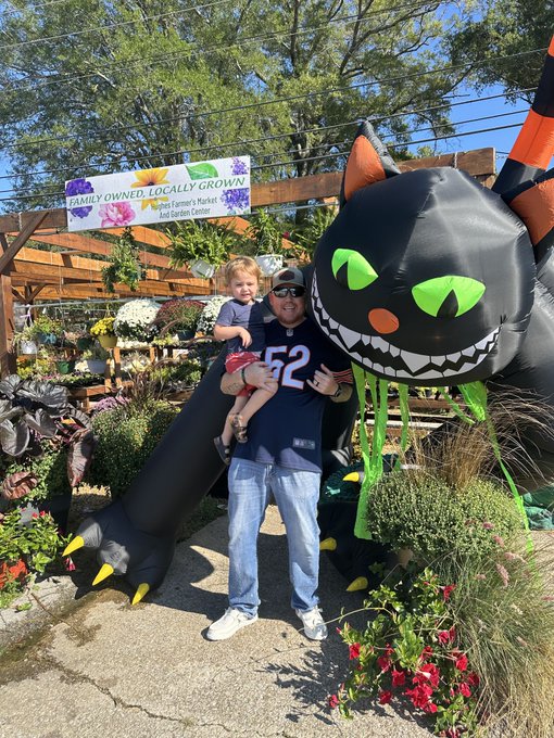 A man wearing a '52' jersey holds a small child while posing in front of a large inflatable black cat decoration at a family-owned garden center.