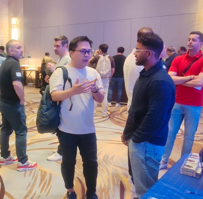 A man in a white shirt gestures with his hands while speaking to another man wearing glasses and a black sweater at what appears to be a networking or conference event.