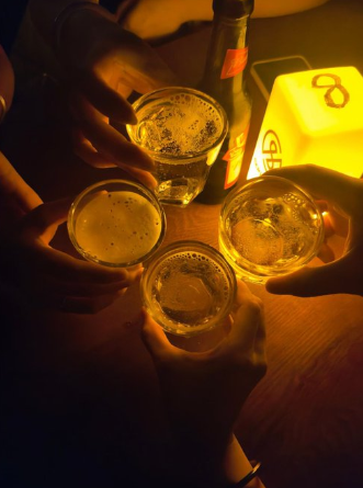Four people clinking glasses of beer in a toast under warm orange lighting, with a glowing lamp visible in the background.