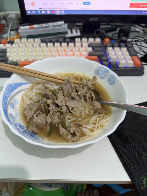 A bowl of beef noodle soup with chopsticks and a spoon resting on the rim, placed on a desk in front of an illuminated keyboard and monitor.