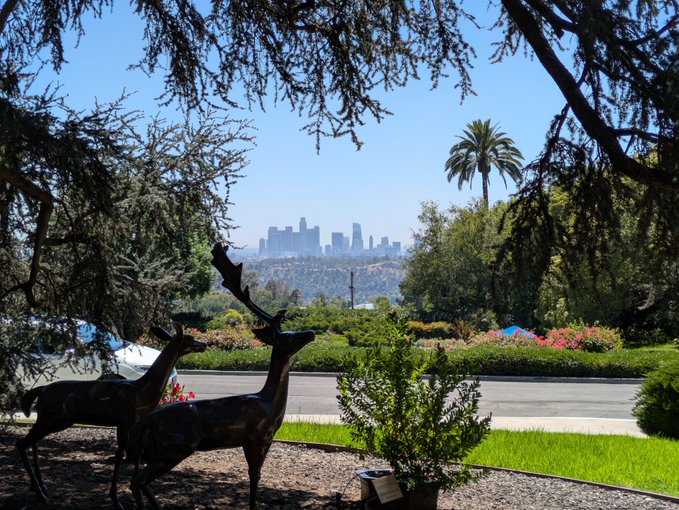 Outdoor scenic view showing two deer sculptures in the foreground with the Los Angeles skyline visible in the distance, framed by trees and a garden with flowers and a palm tree under a clear blue sky.