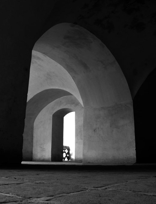 A black and white architectural photograph showing a large arched doorway or passageway with light streaming through a smaller opening in the background.