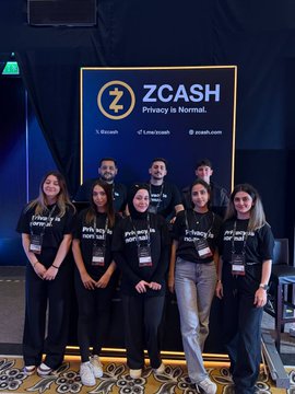 The image features a group of people posing for a photo, all wearing black t-shirts with the words "Privacy Is Normal #Zcash" printed on them. The group consists of at least nine individuals standing close together against a dark background. They are positioned in front of a large screen displaying the Zcash logo and the phrase "Privacy is normal".