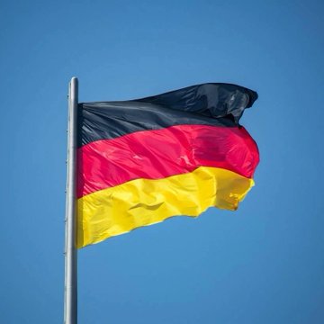 The German national flag with its black, red, and gold horizontal stripes waves against a clear blue sky.