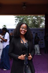A woman with curly dark hair smiling at the camera, wearing a black jacket and jeans, standing on a purple carpet at what appears to be an event.