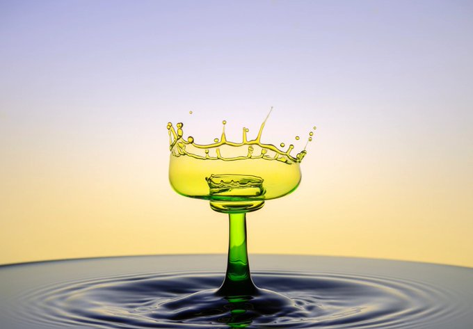 An artistic 3D rendering of a green liquid splashing in a margarita-style glass, suspended above a rippling water surface against a gradient yellow and blue sky.
