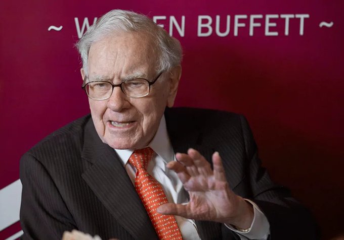 Warren Buffett speaking in front of a red backdrop displaying his name.