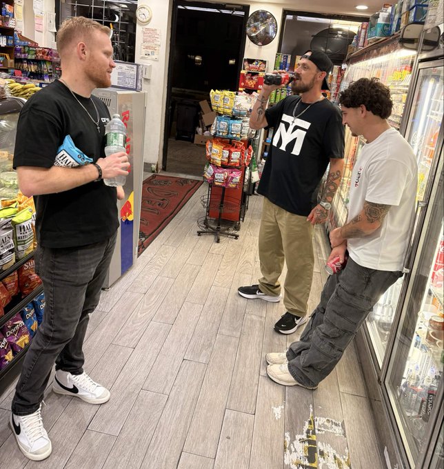 Three young men are standing in the aisle of a convenience store, with one drinking from a bottle of soda, surrounded by shelves stocked with snacks and a refrigerated display case.