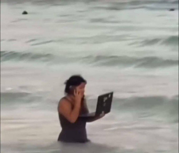 A woman in a dark swimsuit stands waist-deep in the ocean while balancing a laptop in one hand and holding a phone to her ear, multitasking in the water. The image humorously illustrates the post's joke about crypto or meme-stock "runners," depicting how far people are willing to go—literally into the sea—while glued to their screens to catch the next big market spike.