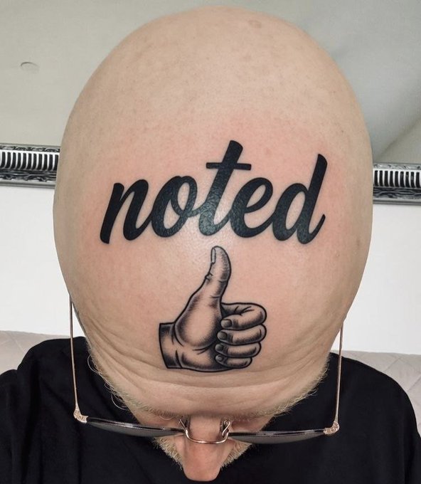 A close-up photo of a bald man's head shows a black ink tattoo featuring the word 'noted' in cursive above a drawing of a hand giving a thumbs-up.