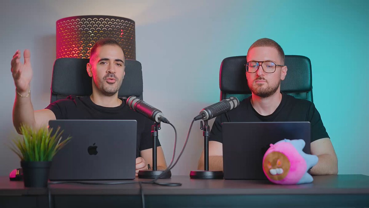 The image shows two men sitting at a table with microphones, each using their own laptop computer. They appear to be engaged in an interview or discussion about cryptocurrency. The background features a blue wall that complements the color of one man's shirt, while another man is wearing glasses and has a pink stuffed animal on his lap. A potted plant can also be seen near them.