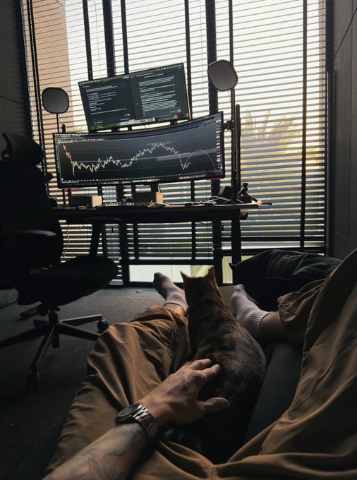 A first-person view of someone relaxing with a cat on their lap, looking toward a dual-monitor trading setup displaying charts and code by a window with blinds.