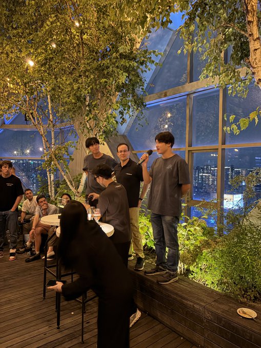A group of men stand holding microphones on an elevated stage at an outdoor rooftop event at night, with string lights in the trees and a city skyline visible through glass windows behind them.