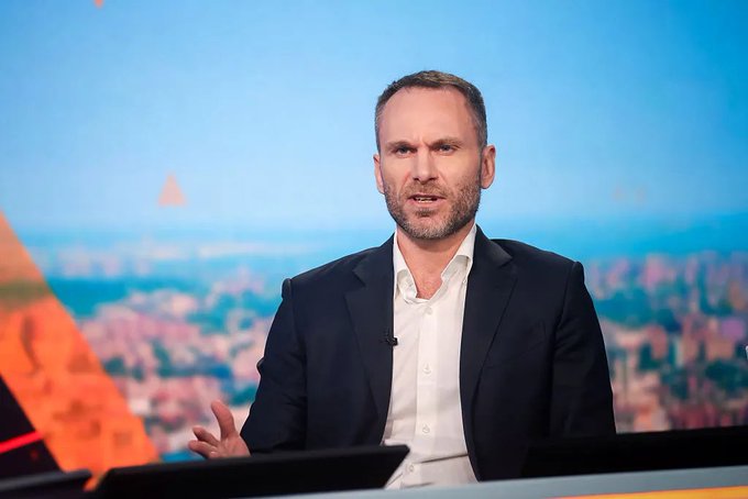 A man in a dark suit and white shirt, likely a presenter or host, speaks while gesturing in a TV studio with a cityscape backdrop, accompanying a discussion on "Tether, Stable Currency and Stable Intelligence."