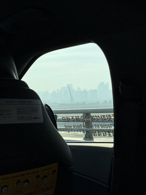 View from inside a vehicle (likely a taxi in Korea) showing a bridge over water with a city skyline in the hazy background; the window has Korean text and 'Closed sale on the 2nd floor.'