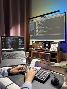A person works at a desk with a laptop, external monitor, and additional displays, typing code on a keyboard in a home office setup.