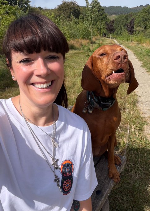 A selfie of a smiling woman with short brown hair wearing a white t-shirt, posing next to a reddish-brown Vizsla dog outdoors in a sunny park setting.
