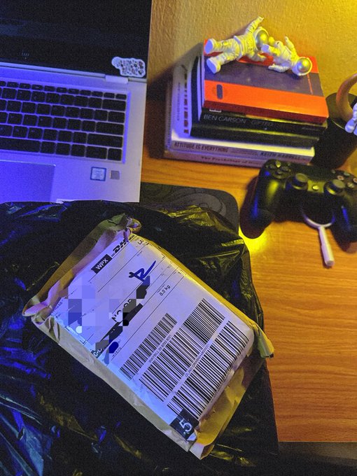 A wooden desk holds a laptop, a stack of books including "KETTLEBALLS EXERCISE" and one by "BEN CARSON", a PS4 controller, an astronaut figurine, and a shipping label on a padded envelope.