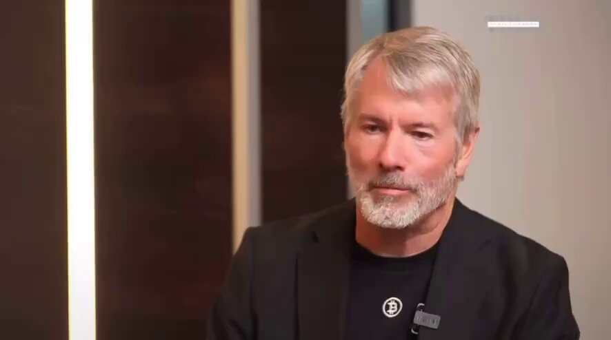 The image features a man wearing a black suit jacket with a white collar and a beard. He has grey hair and appears to be looking directly at the camera, engaging the viewer with his expression. The background of the image shows a wall that is brown in color. There are also some text elements present on this wall, including the phrase "If Strategy sells Bitcoin, they will buy multiples more BTC than they sold" which seems to be related to the post about Michael Saylor and bitcoin.