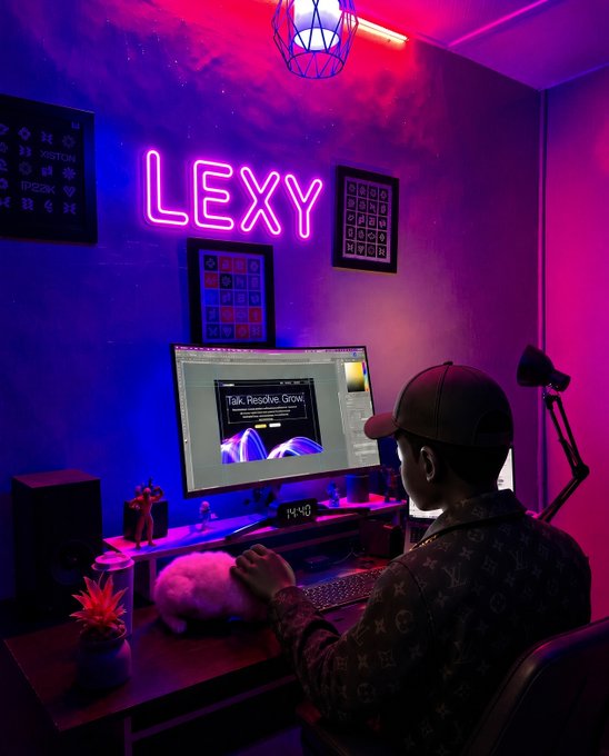 A moody, neon-lit setup of a person wearing a cap working at a computer in a room with a pink 'LEXY' neon sign on the wall and a white cat on the desk.