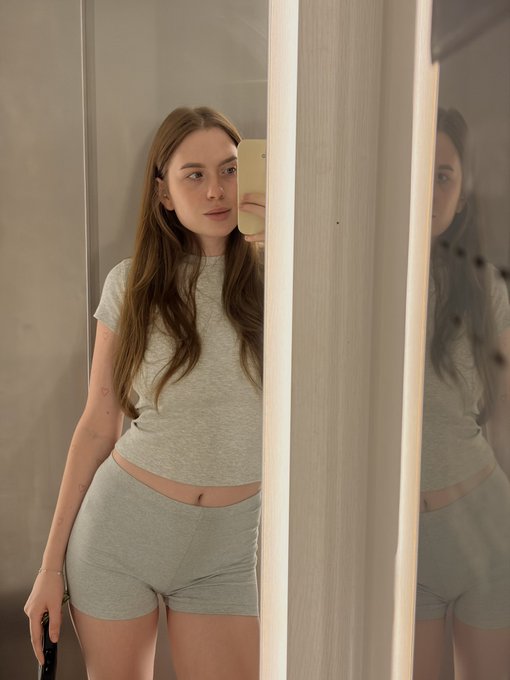 A young woman takes a mirror selfie wearing a grey fitted t-shirt and matching shorts, revealing her midriff.