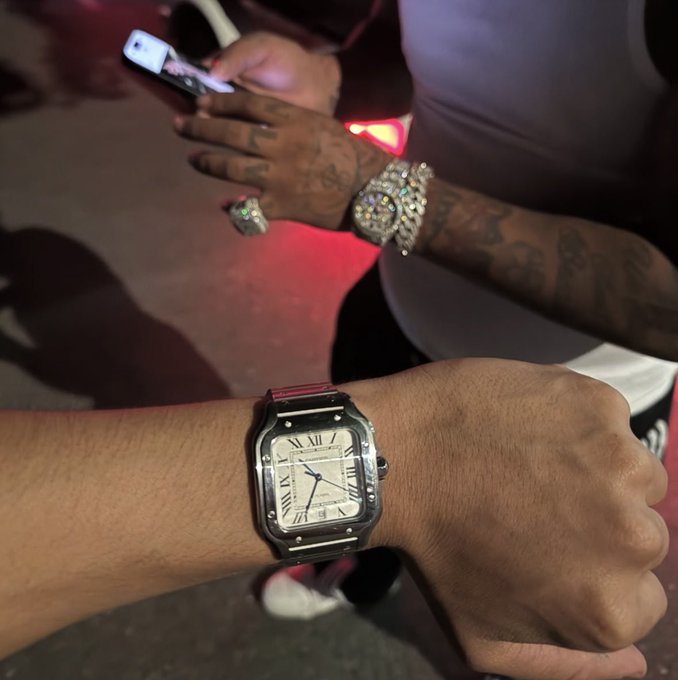 A close-up shows a wrist wearing a Cartier Santos watch with a white dial and Roman numerals, while another tattooed hand in the background holds a phone and sports a diamond-encrusted watch and ring, displaying the kind of luxury flex often used to entice engagement in social media giveaways like the "$110 to a new follower" post.