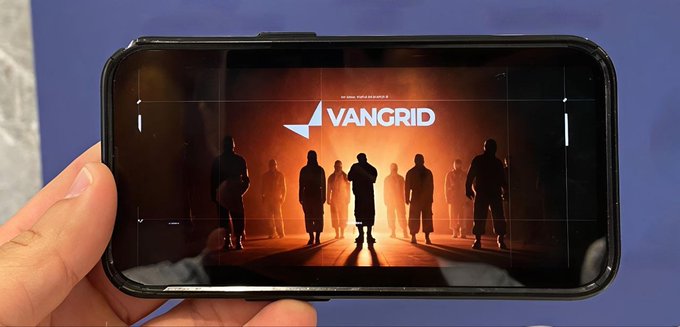 A hand holding a smartphone displaying the VANGRID logo with silhouettes of people against an orange glowing background.