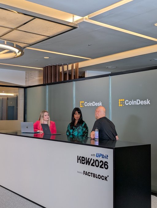 Three people sit at a CoinDesk-branded desk at the KBW2026 conference hosted by FactBlock with Upbit, featuring two women and a bald man in a black shirt.
