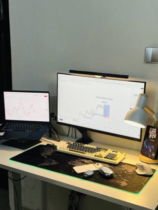 A desk setup featuring a laptop and monitor displaying financial charts, a mechanical keyboard, and a world map mousepad with green LED lighting.