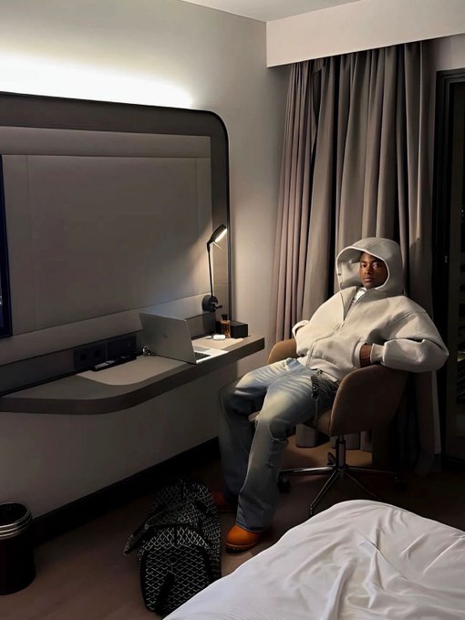 A person wearing a light-colored hoodie sits in an office chair at a desk with a laptop, taking a mirror selfie in a dimly lit hotel room.