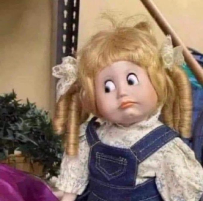 A vintage doll with blonde curly hair in pigtails, wide painted eyes, and an orange mouth, wearing a floral blouse under denim overalls.