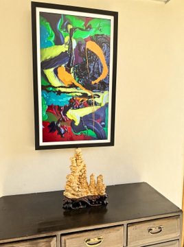 A colorful abstract painting of a toucan hangs above a wooden cabinet displaying a carved stone sculpture.