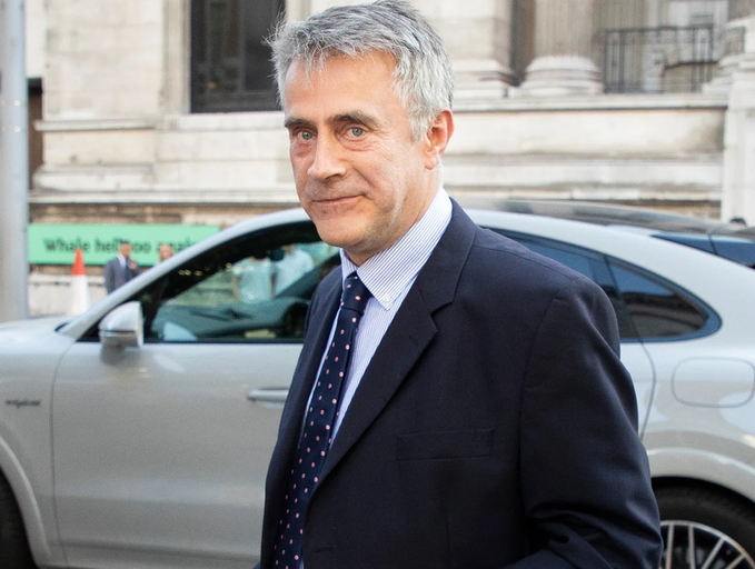A portrait photo of a grey-haired man in a dark suit with a polka dot tie standing in front of a silver Bentley car.